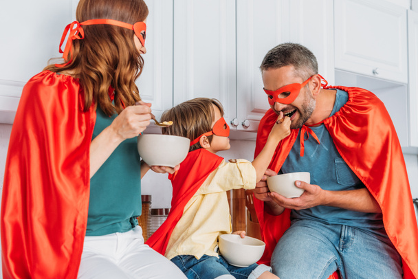 happy family in costumes of superheroes eating breakfast while son feeding father with flakes - Zdjęcie, obraz