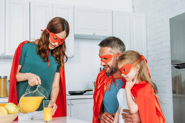 スーパーヒーローの衣装を着た父と娘の近くにガラスにオレンジジュースを注ぐ母親 - 写真・画像