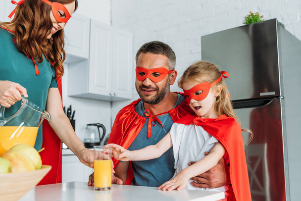 Mutter gießt Orangensaft in Glas neben Ehemann und Tochter in Superheldenkostümen - Foto, Bild