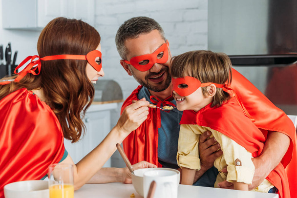mother feeding son with flakes, while family in costumes of superheroes having breakfast - Foto, Imagem