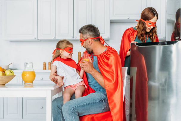 family in costumes of superheroes spending time in kitchen, while father and daughter drinking orange juice and mother standing near fridge - 写真・画像
