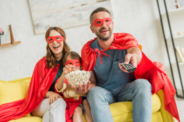 selective focus of father holding remote controller while family in superheroes costumes sitting on sofa with popcorn and watching tv - Fotó, kép