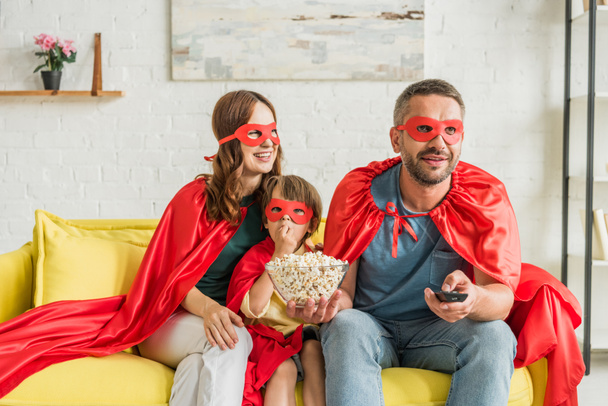 familia alegre en trajes de superhéroes sentados en el sofá y viendo la televisión
 - Foto, imagen
