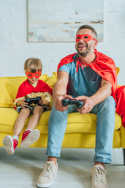 KYIV, UKRAINE - JULY 5, 2019: Cheerful father and son in costumes of superheroes sitting on sofa and playing video game - Photo, Image