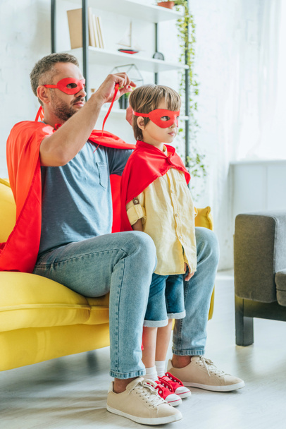 father in superhero costume putting red mask on son while sitting on sofa at home - Photo, Image