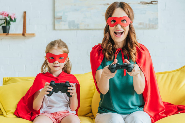 KYIV, UKRAINE - JULY 5, 2019: Cheerful mother and daughter in costumes of superheroes playing video game while sitting on sofa at home - Photo, image
