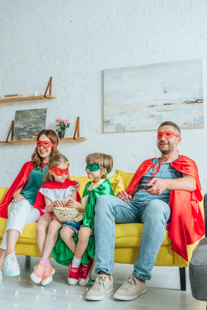parents and two adorable kids in superheroes costumes watching tv while sitting on sofa at home - Foto, Imagem