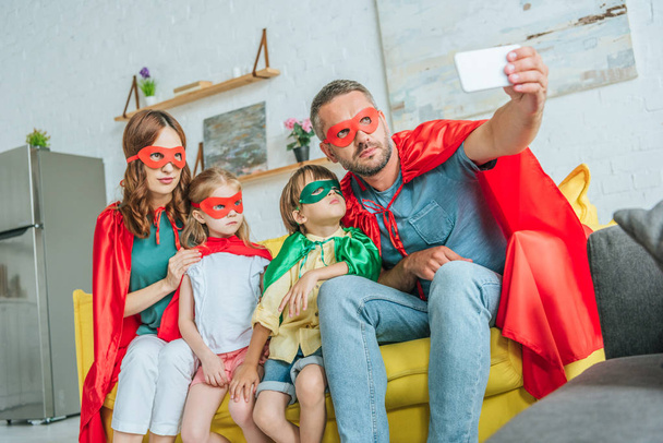 family in costumes of superheroes taking selfie on smartphone while sitting on sofa at home - Foto, immagini
