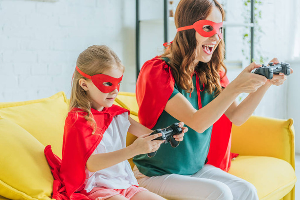 KYIV, UKRAINE - JULY 5, 2019: Excited woman and cute chid in costumes of superheroes playing video game at home - Photo, image