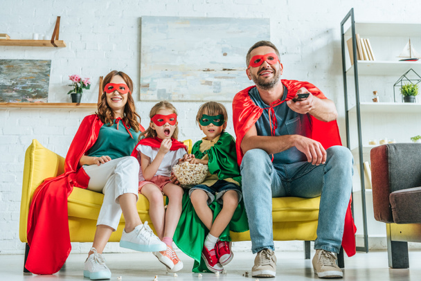 happy family in costumes of superheroes watching tv while sitting on sofa with popcorn - Photo, Image