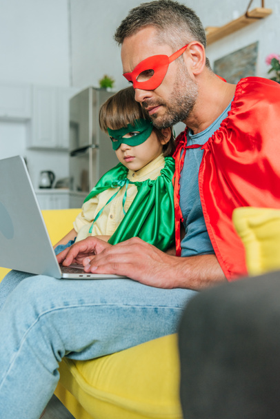 selective focus of attentive father and son in superheroes costumes using laptop at home - Fotografie, Obrázek
