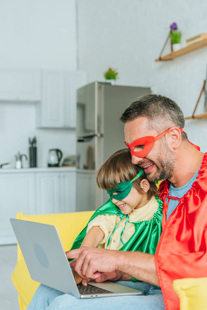 smiling father and son in superheroes costumes using laptop at home - Fotó, kép