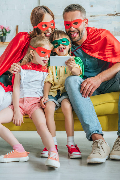 cheerful family in superheroes costumes sticking tongues out while taking selfie on smartphone at home - Foto, afbeelding