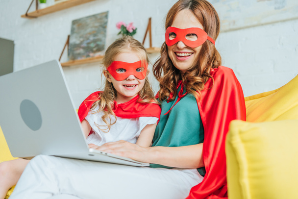 cheerful mom and daughter in costumes of superheroes looking at camera while using laptop at home - Fotoğraf, Görsel