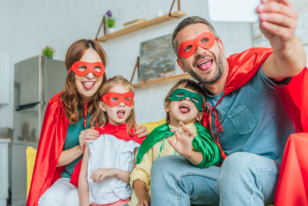 selective focus of happy family in superheroes costumes sticking tongues out while taking selfie on smartphone at home - Фото, зображення