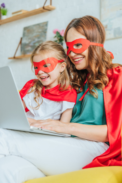 cheerful mom and daughter in costumes of superheroes using laptop at home - Фото, изображение