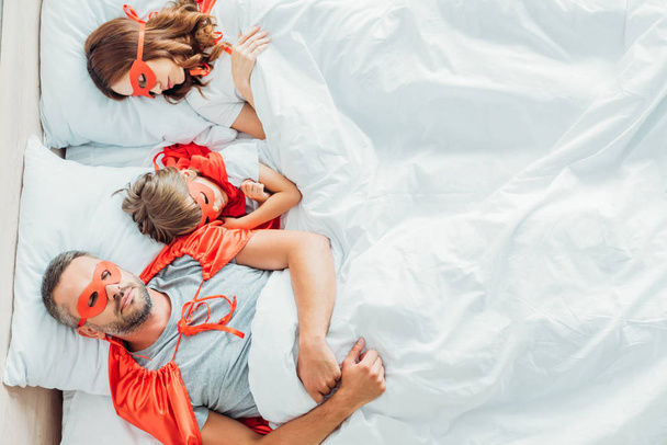 Top uitzicht van vader, moeder en zoon slapen in bed in kostuums van superhelden - Foto, afbeelding