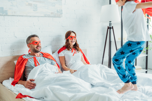 cheerful parents in superheroes costumes looking at son jumping on bed  - Zdjęcie, obraz