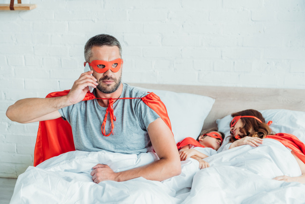 serious man in costume of superhero talking on smartphone while sitting in bed near sleeping wife and son - Photo, Image