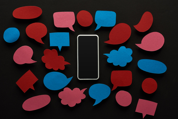 bovenaanzicht van smartphone met leeg scherm op zwarte achtergrond met lege rode en blauwe spraakballonnen, cyberpesten concept - Foto, afbeelding