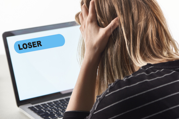 ofendida chica rubia como víctima de cyberbullying sentado cerca de la computadora portátil con mensaje de perdedor en la pantalla
 - Foto, imagen