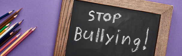 tiro panorâmico de quadro-negro em quadro de madeira com stop bullying lettering perto de lápis coloridos no fundo roxo
 - Foto, Imagem