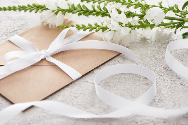 selectieve focus van beige envelop met lint in de buurt van witte bloemen op getextureerde oppervlak   - Foto, afbeelding
