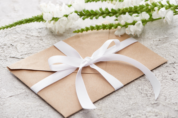 envelope bege com fita branca perto de flores na superfície texturizada cinza
   - Foto, Imagem