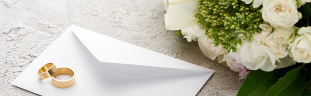 テクスチャ表面上の花の花束の近くに白い封筒上の結婚指輪のパノラマショット  - 写真・画像