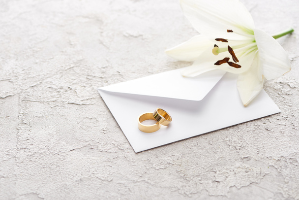 dos anillos de boda de oro en el sobre cerca de lirio blanco en la superficie texturizada
  - Foto, Imagen