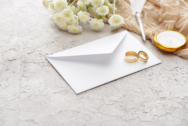 golden rings on white envelope near chrysanthemums, beige sackcloth, quill pen and compass on textured surface  - 写真・画像