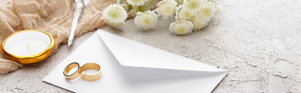 panoramatický záběr zlatých prstenů na bílé obálce blízko chryzantémy, béžové sacké tkaniny, zkosené pero a kompas na texturované ploše  - Fotografie, Obrázek