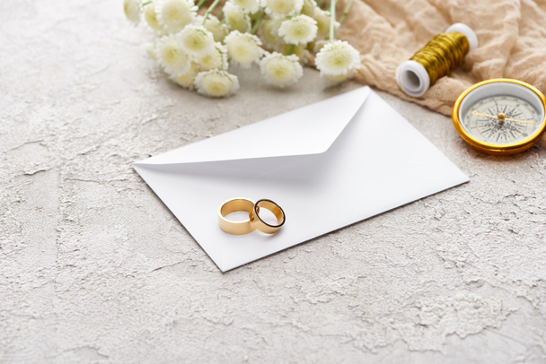 δαχτυλίδια γάμου σε λευκό φάκελο κοντά σε χρυσάνθεμα, μπεζ σάκο, καρούλι και χρυσή πυξίδα σε υφή επιφάνεια  - Φωτογραφία, εικόνα
