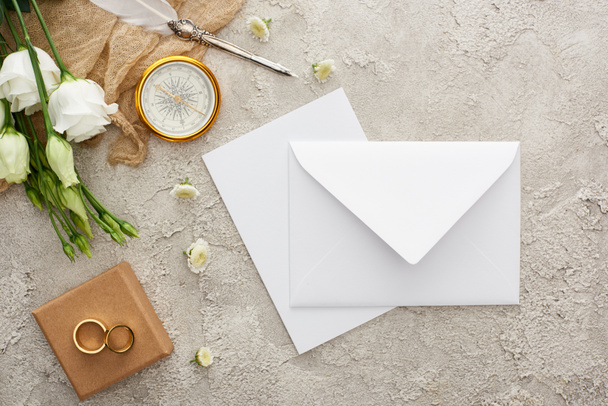 верхний вид белого конверта, компас, обручальные кольца на подарочной коробке на серой текстурированной поверхности
 - Фото, изображение