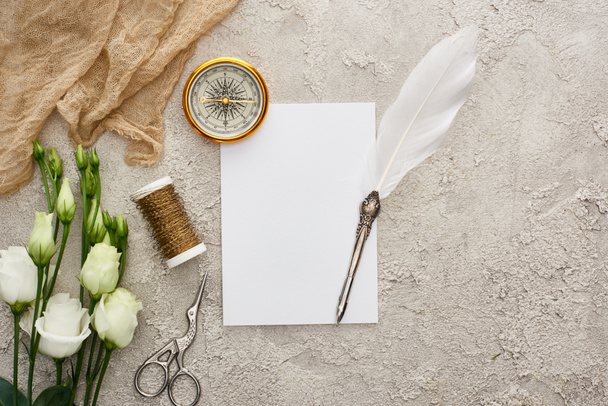 vista superior de pluma pluma de pluma en la tarjeta en blanco cerca de la brújula dorada, saco beige, bobina, tijeras y flores de eustoma blanco en la superficie texturizada gris
 - Foto, Imagen