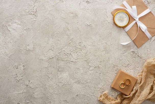 vue du dessus de compas doré, sac beige, bagues dorées sur boîte cadeau et enveloppe avec ruban blanc sur surface texturée
  - Photo, image