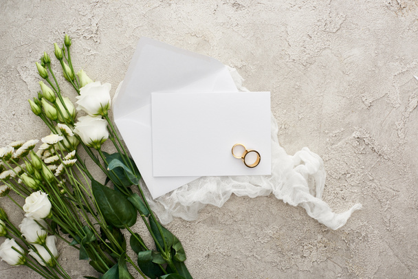 top view of wedding rings on empty card on white cheesecloth near flowers on textured surface - Photo, Image