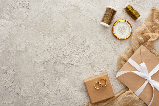 Ansicht von goldenen Ringen auf Geschenkschachtel in der Nähe von Umschlag mit weißem Band in der Nähe von Sacktuch, Spulen und Kompass auf strukturierter Oberfläche  - Foto, Bild