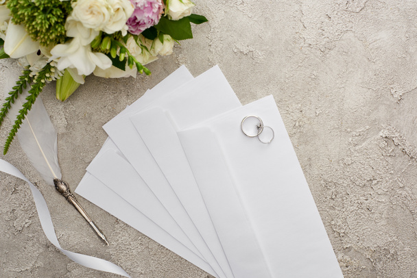 top view of wedding rings on envelopes near quill pen, white ribbon and bouquet on textured surface  - Photo, Image