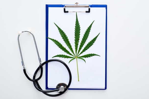 Top zicht op medisch cannabis blad op Klembord nabij stethoscoop op witte achtergrond - Foto, afbeelding