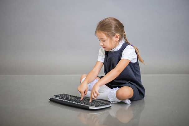 Kız bir okul üniforması içinde birinci sınıf öğrencisi gri bir arka plan üzerinde oturur ve bilgisayardan klavye üzerinde çalışır - Fotoğraf, Görsel