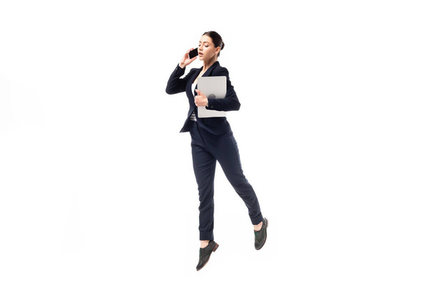 young businesswoman talking on smartphone and holding laptop while dancing isolated on white - Photo, Image