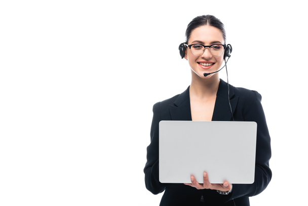 lachende callcenter operator in headset met laptop terwijl je kijkt naar camera geïsoleerd op wit - Foto, afbeelding