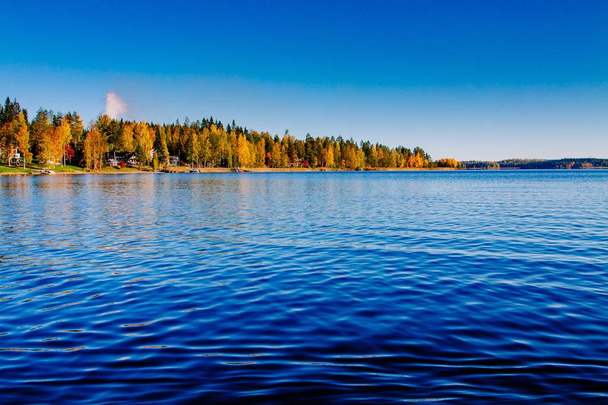 Syksyn lehdet, syksyn värikäs metsä sinisellä järvellä punaisilla mökeillä Suomessa
. - Valokuva, kuva
