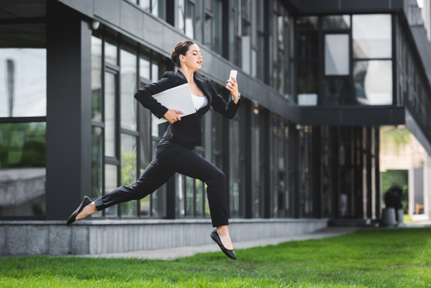 attractive businesswoman levitating while using smartphone and holding laptop - Фото, изображение