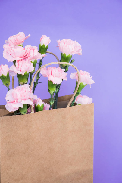 Pink carnation flowers into craft paper bag on purple background. Empty paper bag for your design. Spring`s concept. - Photo, Image