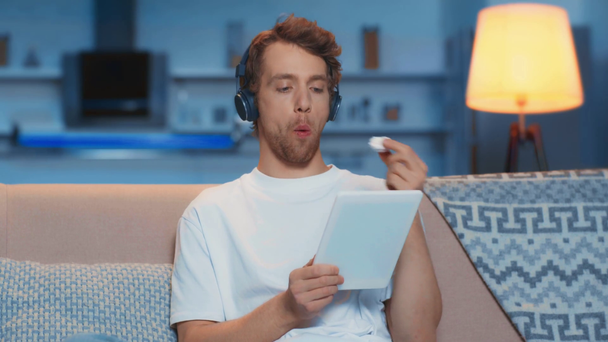 άνθρωπος σε ακουστικά που χρησιμοποιούν ψηφιακό δισκίο και τρώγοντας στο σαλόνι - Πλάνα, βίντεο