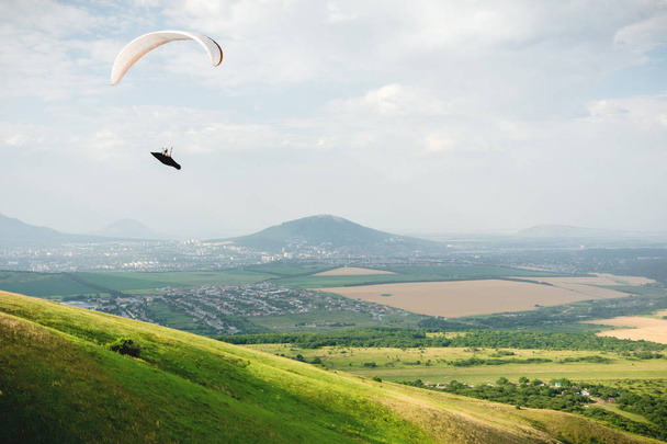 A paraglider flies in the sky in a cocoon suit on a paraglider over the Caucasian countryside with hills and mountains. Paragliding Sport Concept - Фото, изображение