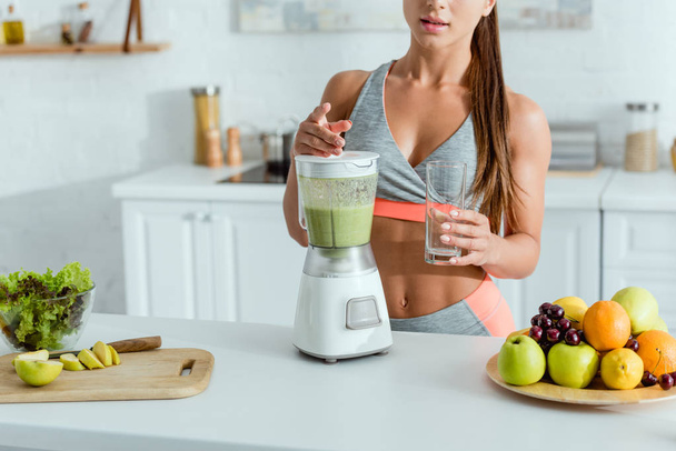 cropped view of sportswoman standing near blender with smoothie and fruits  - Фото, изображение