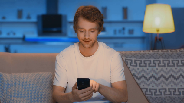 ember használ smartphone és mosolygós nappaliban éjjel - Felvétel, videó